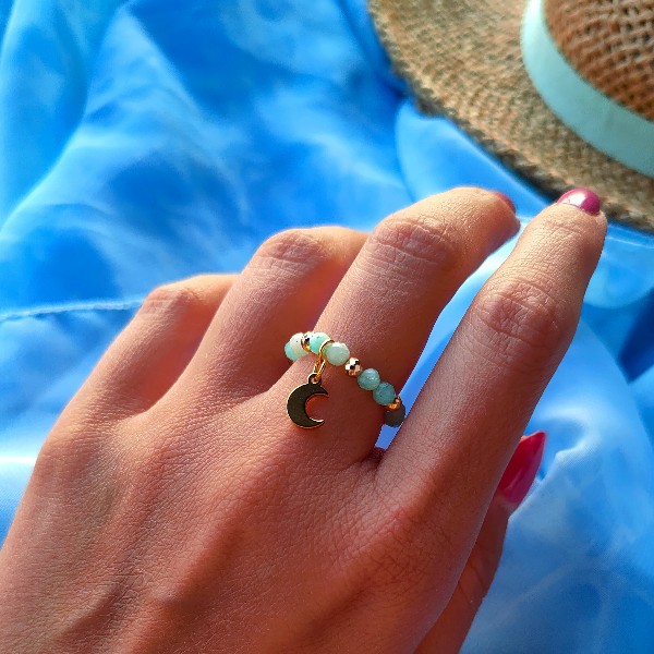 Pierścionek z kolorowymi kamieniami i przywieszką w kształcie księżyca na damskim palcu. W tle kapelusz słomkowy